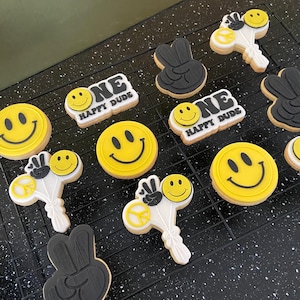 Op de afbeelding: Assortiment van versierde koekjes met een retro thema. Geel en zwarte smiley koekjes, peace teken koekjes en koekjes met de tekst "ONE HAPPY DUDE". De koekjes zijn gerangschikt op een zwart afkoelrek.