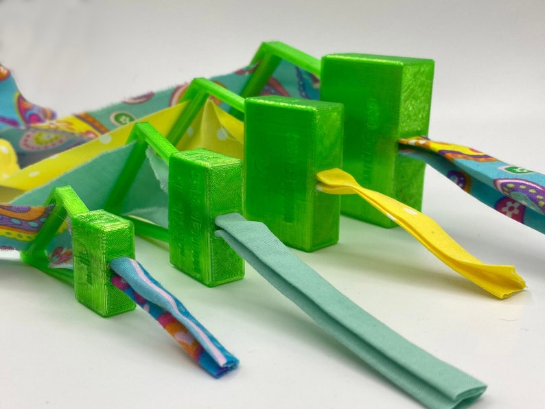 May include: Five green plastic fabric clamps of different sizes, holding colorful fabric strips. The clamps are designed to hold fabric in place while sewing.