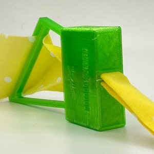 May include: A green 3D printed fabric fastener with a yellow fabric strip threaded through it. The fabric strip has white polka dots.