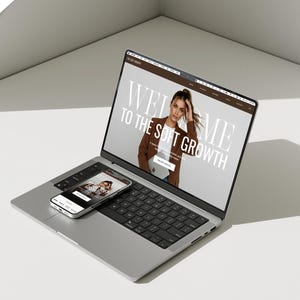 May include: A silver laptop and smartphone displaying a website with the text "WELCOME TO THE SOFT GROWTH". The laptop screen shows a woman in a brown blazer. The smartphone shows a similar image. The devices are on a white surface.