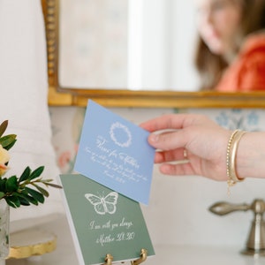 May include: Two small, decorative, square-shaped signs with inspirational messages. The top sign is light blue with a wreath and the text "Wishes for Mother" and a Bible verse. The bottom sign is green with a butterfly and the text "I am with you always. Matthew 28:20". The signs are on a gold metal stand.