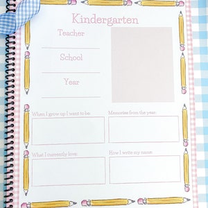 May include: A pink and white gingham patterned journal with a blue ribbon. The cover has a yellow pencil border and the text "Kindergarten" in pink. The journal has blank pages for writing memories from the year.