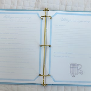 May include: A blue and white baby memory book with the text "While you were growing" on the cover. The book has a binder style with gold rings. The inside pages have lines for writing memories.