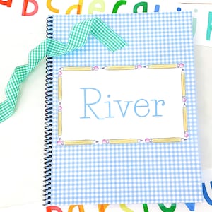 May include: A blue and white gingham notebook with a spiral binding. The cover has a white border with a pencil border and the name "River" written in blue.