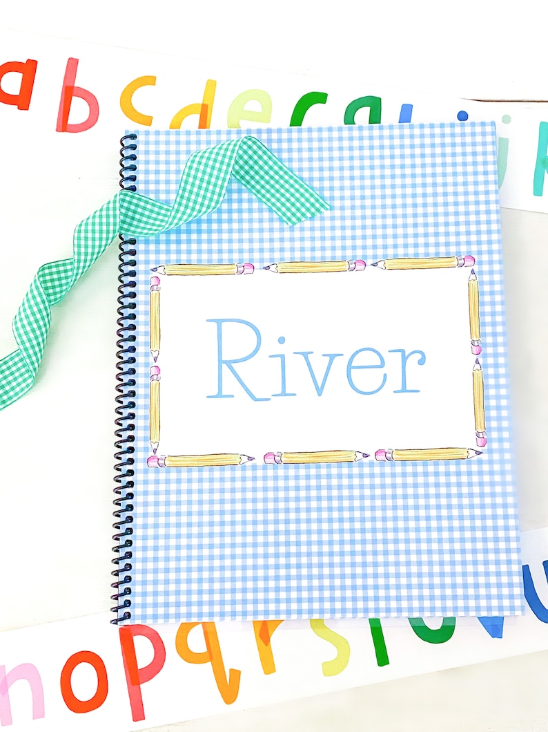 May include: A blue and white gingham notebook with a spiral binding. The cover has a white border with a pencil border and the name "River" written in blue.