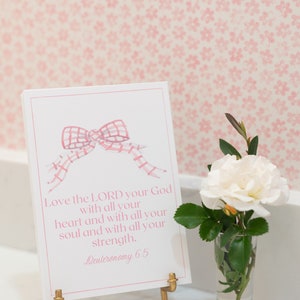 May include: A white card with pink text and a pink and white gingham bow on a gold stand. The text reads "Love the LORD your God with all your heart and with all your soul and with all your strength. Deuteronomy 6.5". A white rose with green leaves is in a clear glass vase.