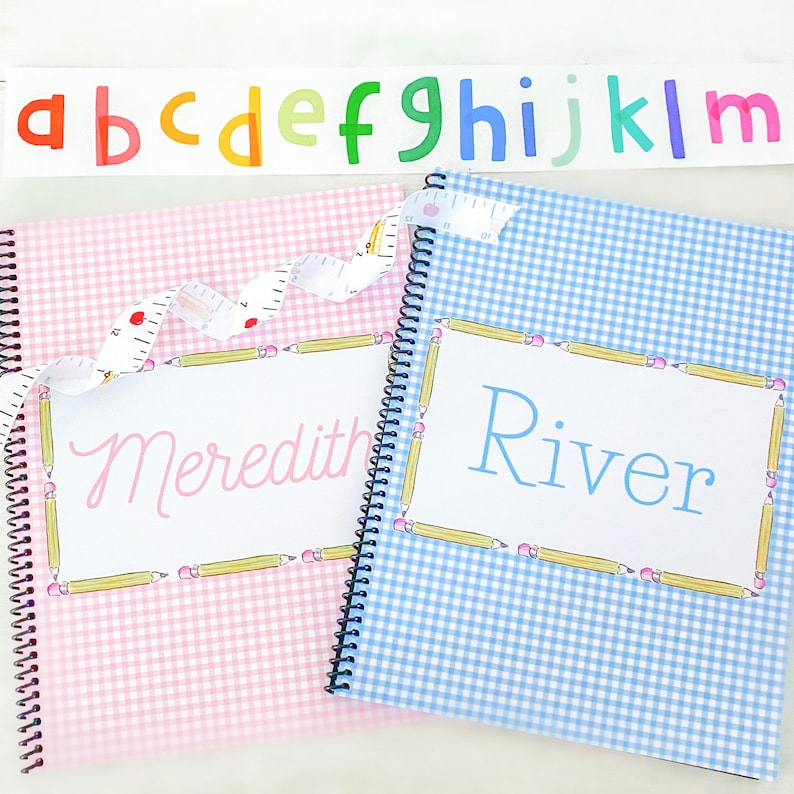 May include: Two spiral bound notebooks with gingham patterns. The pink notebook has the name Meredith written on it and the blue notebook has the name River written on it.