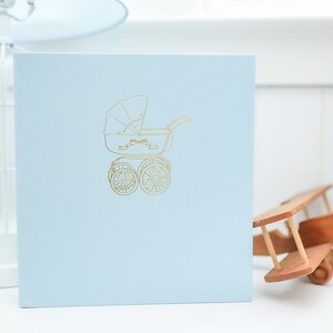 May include: A light blue baby photo album with a gold embossed image of a baby carriage on the front cover.