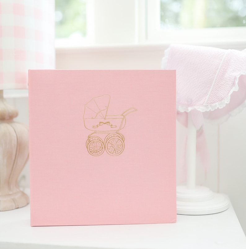May include: A pink photo album with a gold outline of a baby carriage on the front.