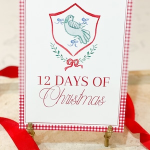 May include: A white card with a red gingham border. The card features a watercolor illustration of a dove with a red bow and green leaves. The text "12 Days of Christmas" is printed in red.