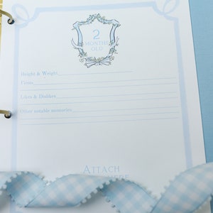 May include: A blue and white baby memory book page with a decorative border and a crest that says "2 Months Old". The page has lines for recording height, weight, firsts, likes and dislikes, and other notable memories.