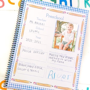 May include: A blue and white checkered preschool memory book with a photo of a young boy. The book has a title that says "Preschool" and includes spaces to fill in information about the child's teacher, school, year, what they want to be when they grow up, what they love, and memories from the year. The book also includes a space for the child to write their name.