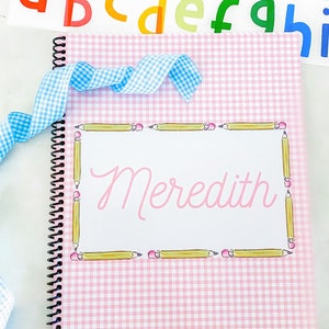May include: A pink and white gingham notebook with a spiral binding. The cover has a yellow pencil border and the name "Meredith" written in pink inside the border.