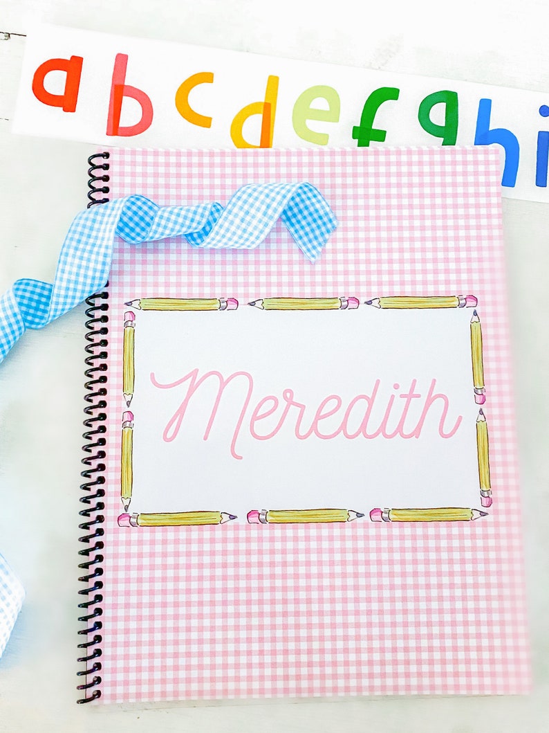 May include: A pink and white gingham notebook with a spiral binding. The cover has a yellow pencil border and the name "Meredith" written in pink inside the border.