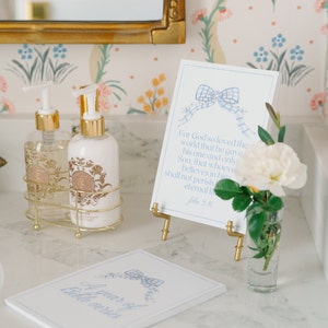 May include: A white card with a blue ribbon and the text "For God so loved the world that he gave his one and only Son, that whoever believes in him shall not perish but have eternal life. John 3:16" on a gold stand. Two bottles of lotion with gold caps and labels are on a gold wire rack. A small glass vase with a white flower is on a white marble surface. A white card with the text "A year of Bible verses" is on the surface.