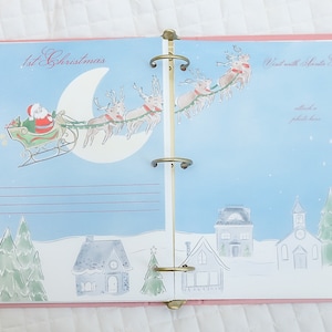 May include: A pink and blue Christmas memory book with a page featuring a Santa Claus illustration flying in a sleigh pulled by reindeer. The page has a title that reads "1st Christmas" and a space for a photo.