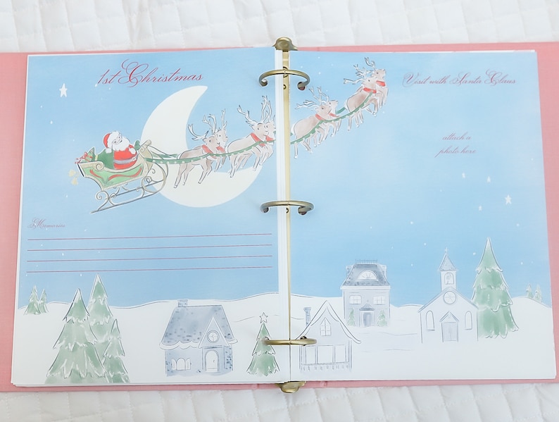 May include: A pink and blue Christmas memory book with a page featuring a Santa Claus illustration flying in a sleigh pulled by reindeer. The page has a title that reads "1st Christmas" and a space for a photo.