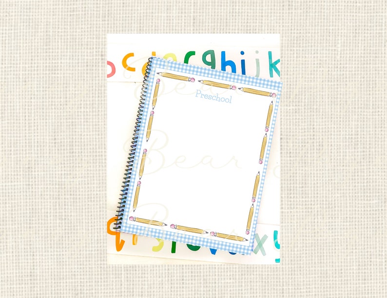 May include: A blue and white checkered notebook with a pencil border. The notebook says "Preschool" on the front. The notebook is on top of a sheet of paper with colorful letters.