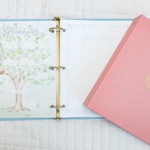 May include: A pink binder with a gold design of a baby carriage on the front. The binder is open to a page with a watercolor illustration of a family tree and a blank page for writing.