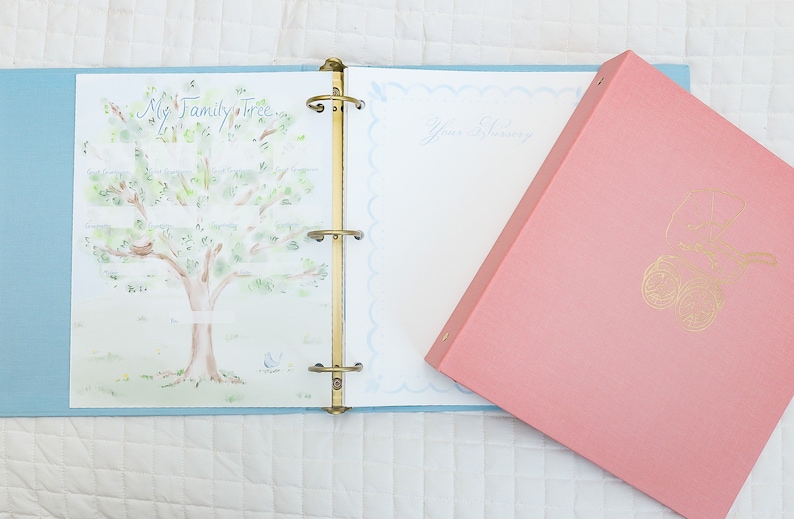 May include: A pink binder with a gold design of a baby carriage on the front. The binder is open to a page with a watercolor illustration of a family tree and a blank page for writing.