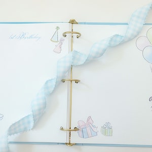 May include: A blue and white scrapbook with a gold binder. The cover has a drawing of a cupcake with a candle, a party hat, and balloons. The text "1st Birthday" is written on the cover.