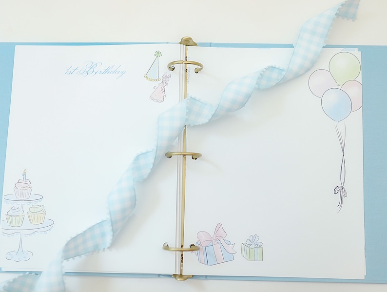 May include: A blue and white scrapbook with a gold binder. The cover has a drawing of a cupcake with a candle, a party hat, and balloons. The text "1st Birthday" is written on the cover.