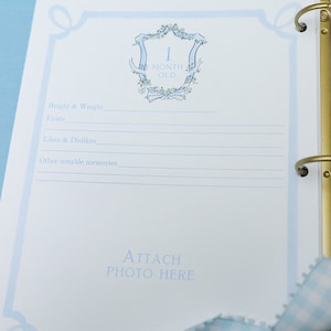 May include: A blue and white baby memory book with a decorative floral design and the text "1 Month Old". The book has a place to attach a photo and record baby's height, weight, firsts, likes and dislikes, and other notable memories.