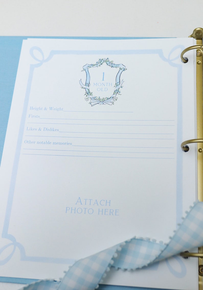 May include: A blue and white baby memory book with a decorative floral design and the text "1 Month Old". The book has a place to attach a photo and record baby's height, weight, firsts, likes and dislikes, and other notable memories.