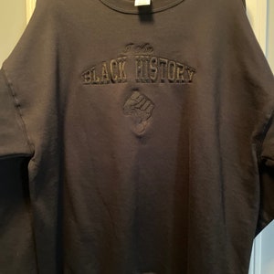 May include: Black sweatshirt with the embroidered text "I Am BLACK HISTORY" in a stylized font. Below the text is an embroidered fist. The sweatshirt is hanging on a black hanger.