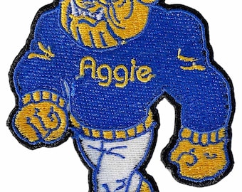 May include: Embroidered patch featuring a stylized bulldog mascot in blue and gold. The bulldog is wearing a blue sweater with the word "Aggie" in gold lettering. The patch has a black border and is designed for application on clothing or accessories.