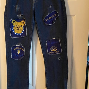 May include: Blue denim jeans with patches. The patches include a bulldog, a crown, and text that says "Aggie Born Aggie Bred" and "Aggie Alum".