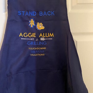 May include: A navy blue apron with a pocket. It has the text "Stand Back Aggie Alum is Grilling Touchdowns Tailgates Traditions" embroidered on it.