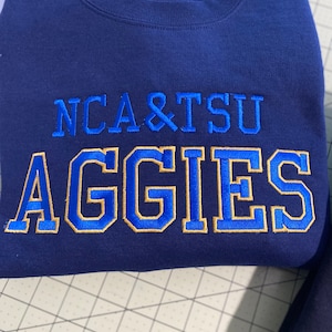 May include: Navy blue sweatshirt with gold and blue embroidered text that reads "NCA&TSU Aggies".
