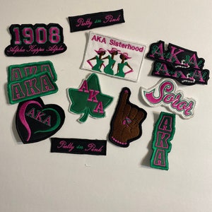 May include: A collection of iron-on patches featuring the letters AKA in various styles and colors, including green, pink, and black. Some patches include the words "Pretty in Pink", "AKA Sisterhood", and "Soror".