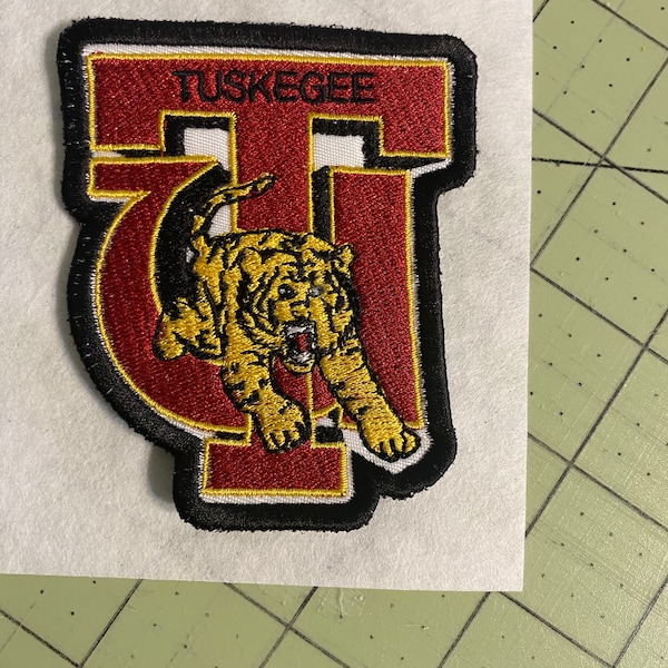 Op de afbeelding: Een geborduurde patch in rood en goud met het woord "TUSKEGEE" en een gele tijger in een zwart omlijnde "T".