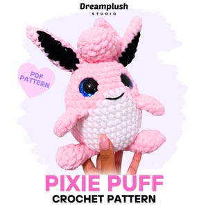 Peut inclure: Un jouet en peluche crocheté rose et blanc avec de grands yeux noirs et bleus, des oreilles pointues avec des sections intérieures noires et une touffe de laine sur le dessus. Le texte indique Dreamplush Studio, PDF Pattern, Pixie Puff Crochet Pattern.