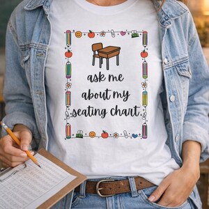 Puede incluir: Camiseta blanca con el texto "ask me about my seating chart" en negro. La camiseta tiene un borde decorativo con lápices, manzanas y otros gráficos escolares. La persona lleva una chaqueta vaquera y vaqueros azules.