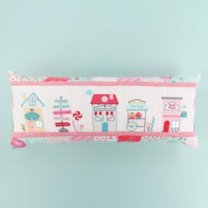 May include: A rectangular decorative pillow featuring a whimsical town scene with buildings, signs, and candy. The pillow has a white background with pink and blue accents. The buildings include a bakery, a soda shop, and a cupcake stand.