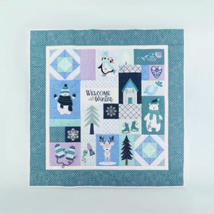 May include: A winter-themed quilt with a blue patterned border. The quilt features various winter-related images, including a snowman, penguin, bear, and deer. The words "Welcome Winter" are also displayed on the quilt.