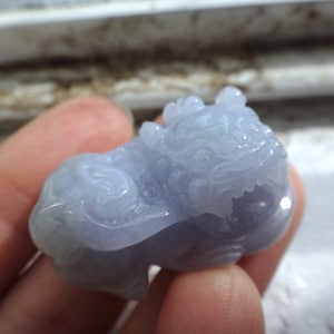 Może przedstawiać: Rzeźba z jasnoniebieskiego jadeitu przedstawiająca mityczną istotę, prawdopodobnie lwa lub smoka, z misternymi detalami. Rzeźba jest gładka i wypolerowana, o delikatnym, prawie przezroczystym wyglądzie.