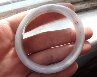May include: A pale lavender jade bangle bracelet with a smooth, polished finish. The bracelet is slightly translucent and has a few small, dark spots.