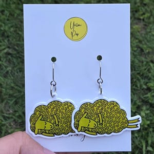 May include: A pair of dangle earrings featuring a yellow sheep graphic with curly wool, attached to silver-toned metal hooks. The earrings are displayed on a white rectangular card with a yellow circular logo containing the text Yellow Pop.