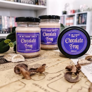 May include: Three glass jars and a candle tin with black lids, labeled "Chocolate Frog" in purple and white. The jars and tin sit on a vintage paper surface, with decorative elements. The candles are a light brown color.