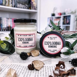 May include: Two candles, one in a glass jar and one in a black tin, with the text "EXPLODING BONBONS" and "COCOA + COCONUT DYNAMITE". The candles are surrounded by decorative elements, including dried botanicals and greenery.