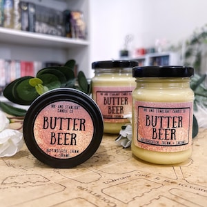 May include: An assortment of Butter Beer scented candles and a tin from Ink and Starlight Candle Co. The candles are in glass jars with black lids, and the tin is black. The labels read "Butter Beer, Butterscotch Cream + Caramel."