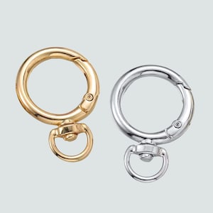 May include: Two metal swivel clasps, one gold-toned and one silver-toned, are displayed against a light gray background. Each clasp features a circular ring with a spring-loaded opening and a smaller, attached ring.