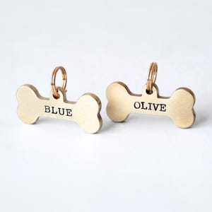 May include: Two bone-shaped pet tags with gold-colored metal. One tag reads "BLUE" and the other reads "OLIVE" in black text. Each tag has a small gold ring at the top for attachment to a collar. The tags are on a white background.