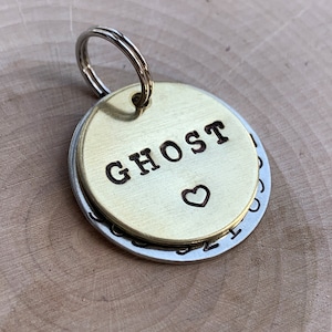 May include: A circular brass pet tag featuring the stamped text GHOST and a small heart symbol, layered over a larger silver-toned circular metal disc. The two metal tags are connected by a silver-colored metal split ring for attachment.