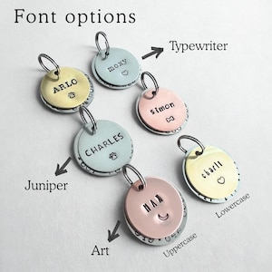May include: Six circular metal pet ID tags in gold, silver, and copper tones, each featuring a split ring. The tags are engraved with names like ARLO, moxy, simon, CHARLES, charli, and MAX, alongside symbols including a paw print, heart, and bone.