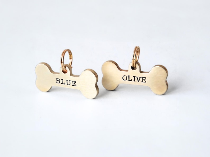 K&ouml;nnte beinhalten: Zwei knochenf&ouml;rmige Haustieranh&auml;nger aus goldfarbenem Metall. Auf einem Anh&auml;nger steht "BLUE" und auf dem anderen "OLIVE" in schwarzer Schrift. Jeder Anh&auml;nger hat einen kleinen goldenen Ring oben zur Befestigung am Halsband. Die Anh&auml;nger befinden sich auf wei&szlig;em Hintergrund.